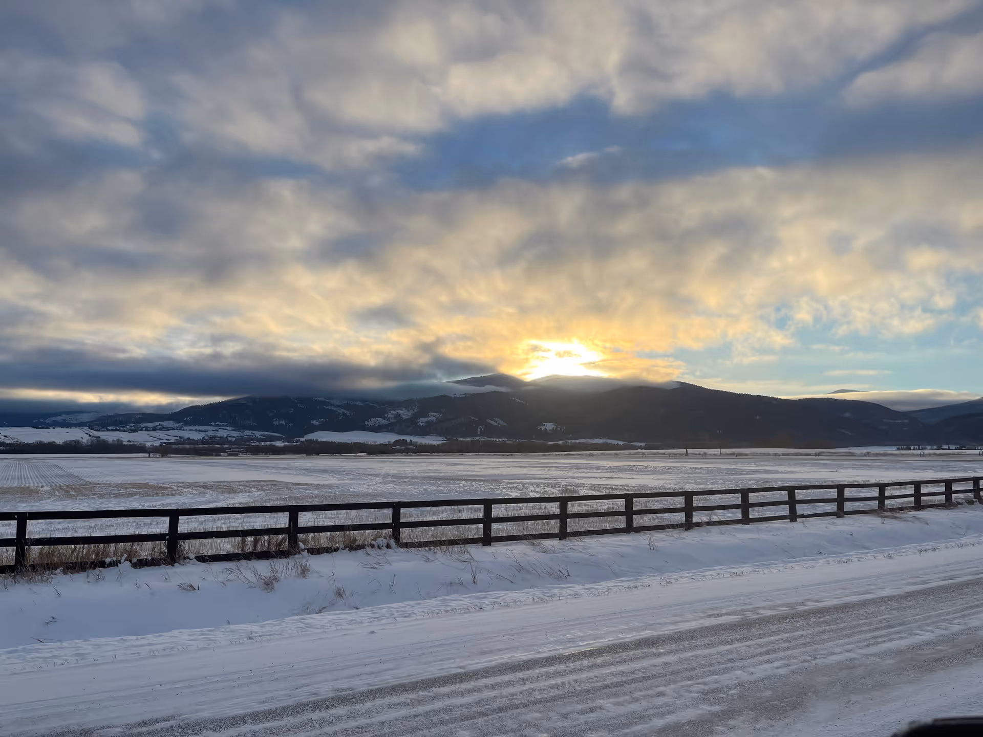 Snow-covered field with a wooden fence in the foreground and mountains in the background under a cloudy sky with the sun setting behind the mountains.