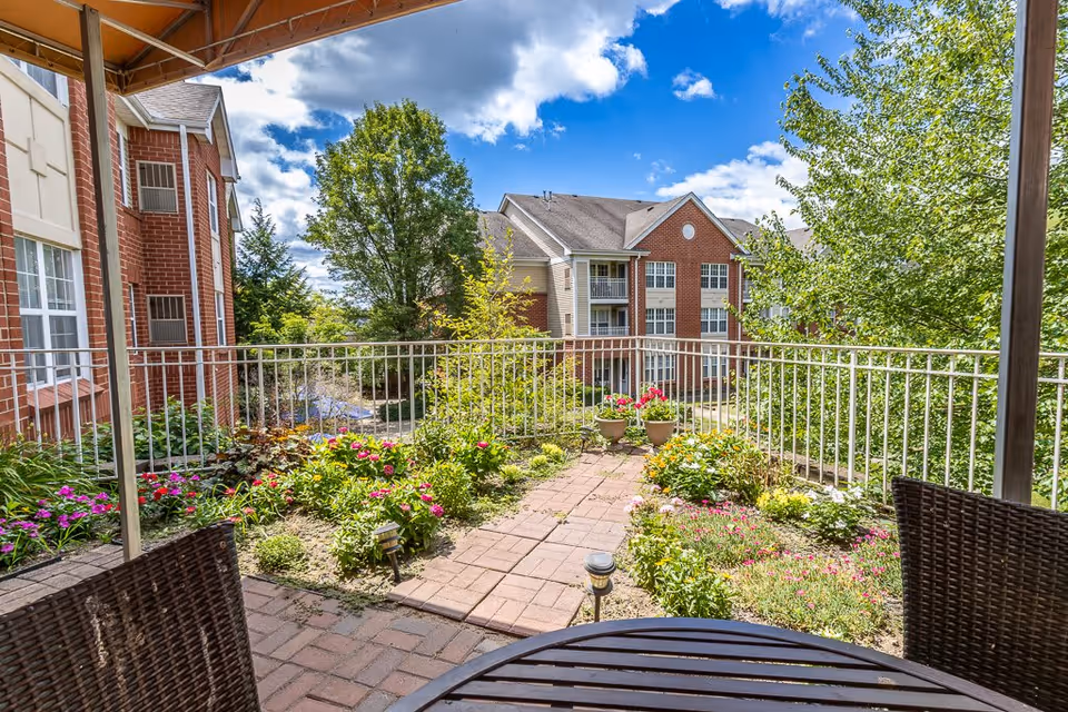 View from a covered patio area with a round table and wicker chairs overlooking a garden with colorful flowers and greenery. In the background, there are multi-story brick and siding residential buildings under a partly cloudy blue sky.