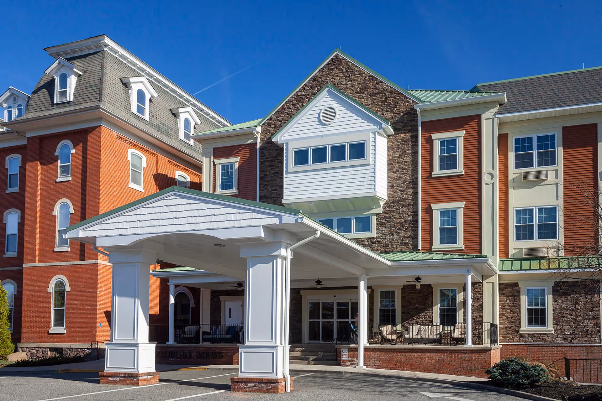 Exterior view of Woodbury Mews senior living facility showing a multi-story building with a covered entrance, brick and stone facade, and multiple windows under a clear blue sky.