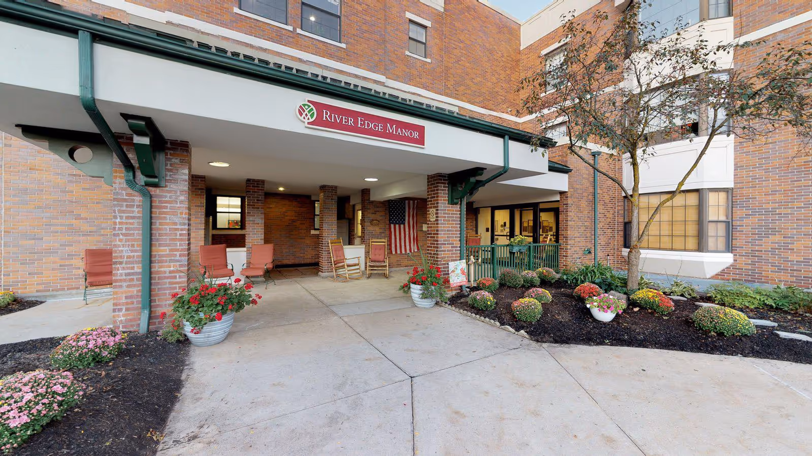 Entrance to River Edge Manor, a senior living facility, showing a covered porch area with several red chairs and rocking chairs. There are flower pots with red and pink flowers on either side of the entrance. The building is made of brick, and there is a small garden area with shrubs and a tree to the right. An American flag is hanging near the entrance.