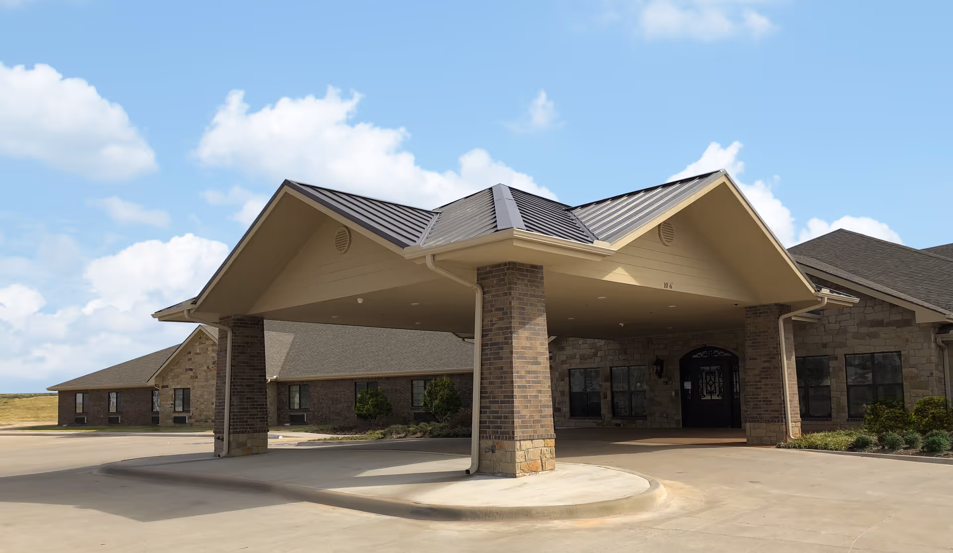 Front exterior view of Promenade Health And Rehabilitation building with a covered entrance supported by brick pillars, surrounded by a paved driveway and landscaped greenery under a partly cloudy sky.