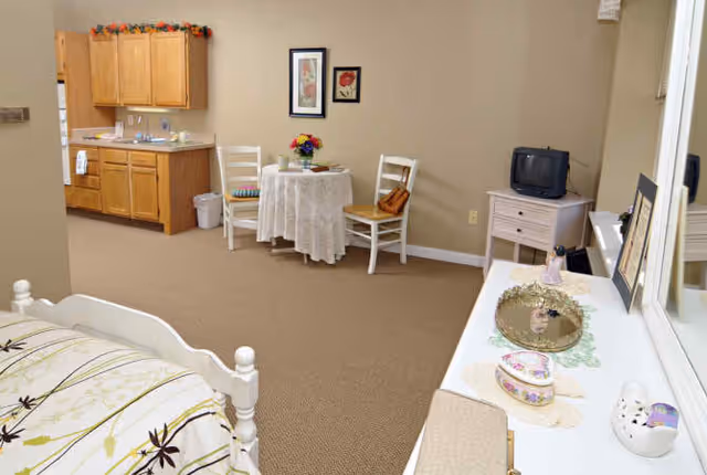 Studio-style interior showing a bed in the foreground, a small kitchenette, a round table with two chairs, and a dresser with a TV and vanity.