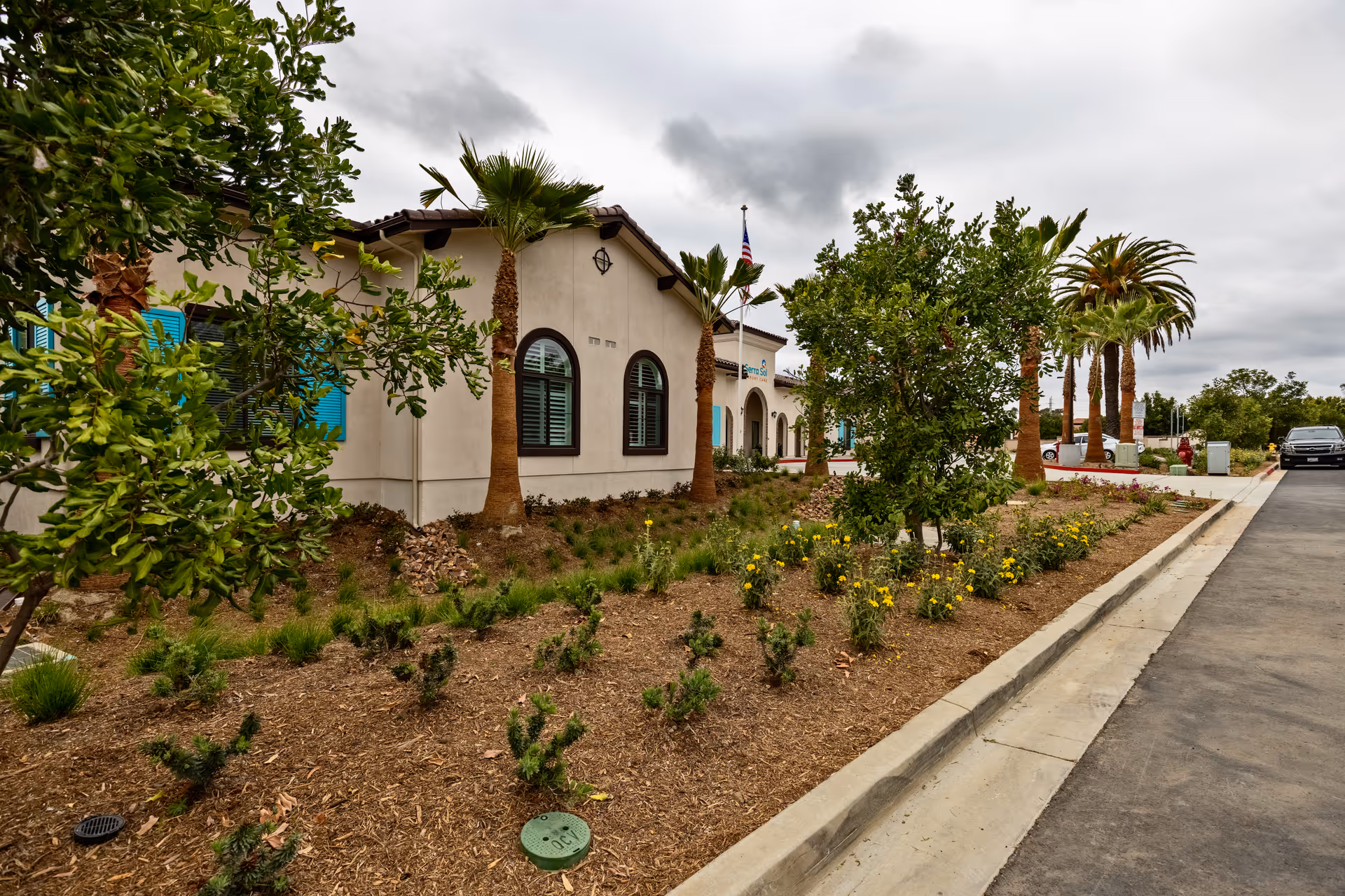 Front exterior of a single-story stucco building with arched windows, palm trees and landscaped planting beds along a driveway under a cloudy sky.