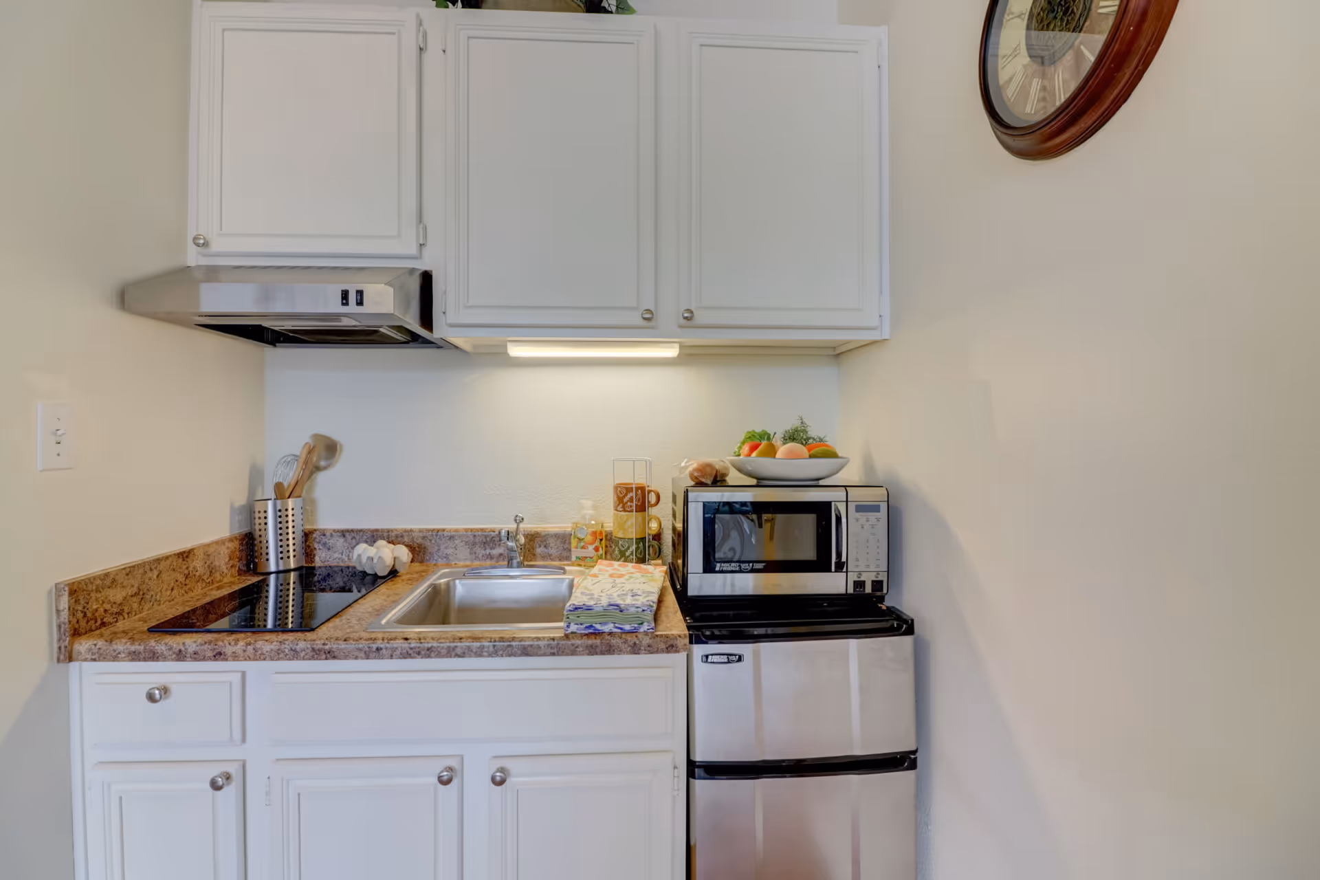 A small kitchen area with white cabinets, a stainless steel sink, a stovetop, a microwave on top of a mini refrigerator, and a bowl of fruit on the microwave. There is a clock on the wall to the right and kitchen utensils in a holder next to the stovetop.