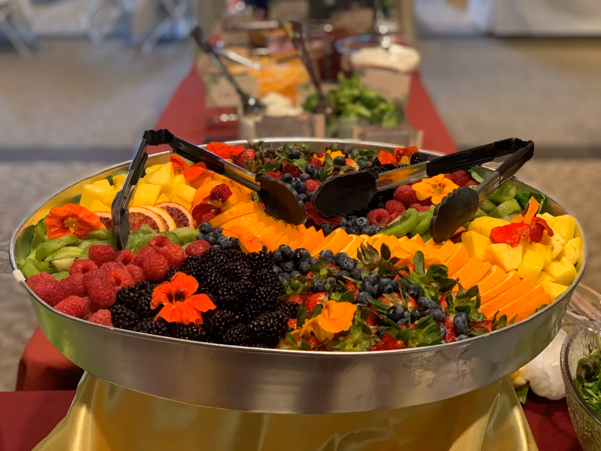 A large round metal tray filled with an assortment of fresh fruits including raspberries, blackberries, blueberries, pineapple chunks, kiwi slices, orange slices, and strawberries, garnished with edible orange flowers. Several black serving tongs are placed on the tray. The tray is set on a table covered with a red and gold tablecloth, with more food trays visible in the blurred background.