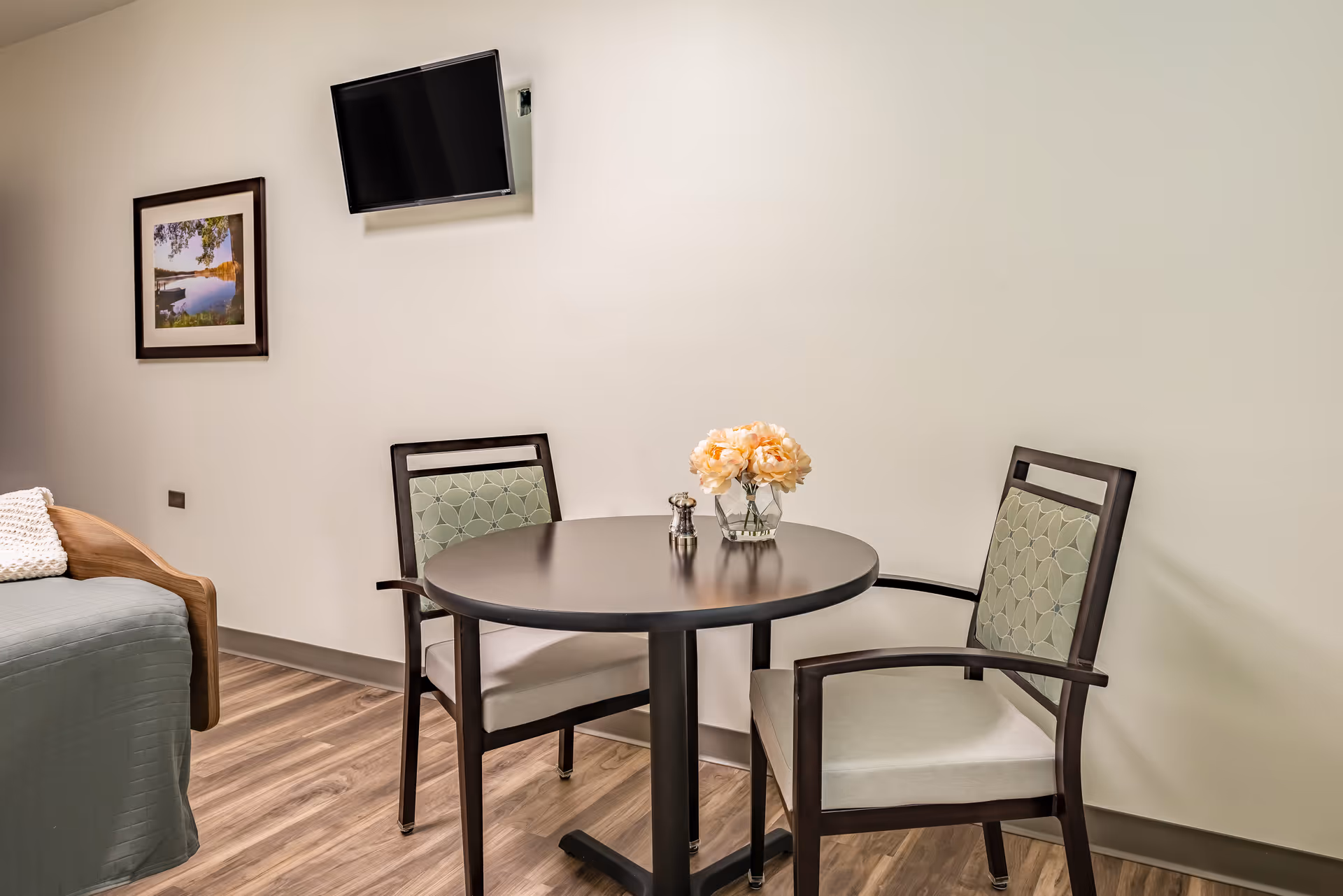 Small resident room with a round table and two chairs, a wall-mounted TV, framed artwork, and part of a bed.