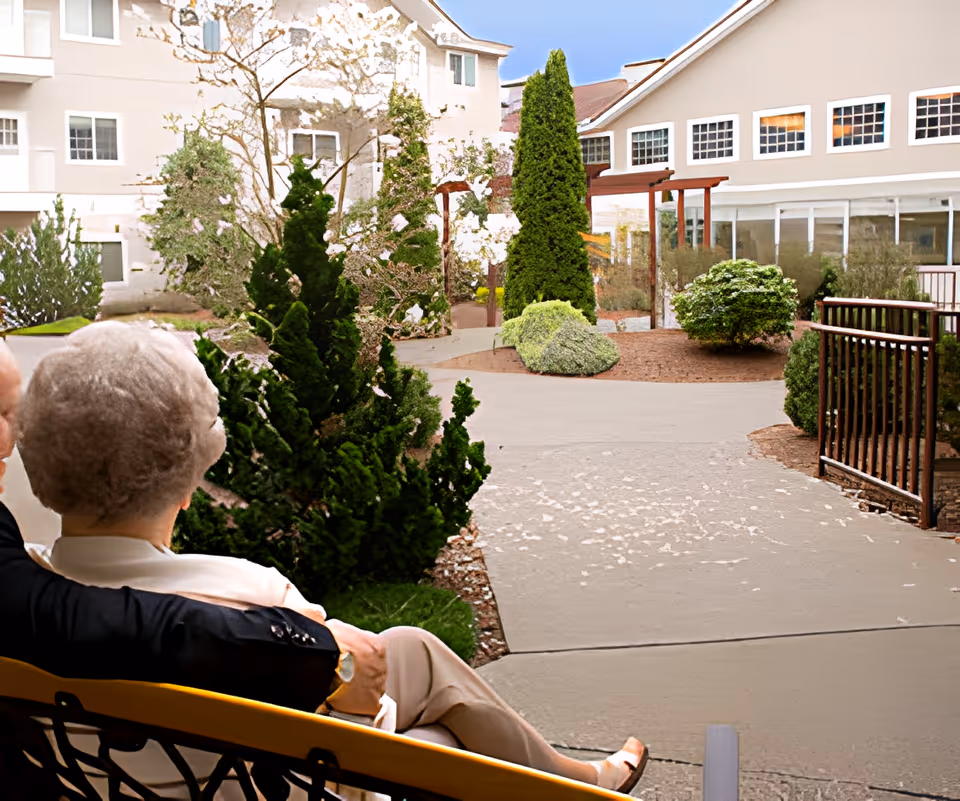 An elderly couple sitting on a bench, looking out at a landscaped outdoor courtyard area with trees, bushes, and a pathway surrounded by buildings.