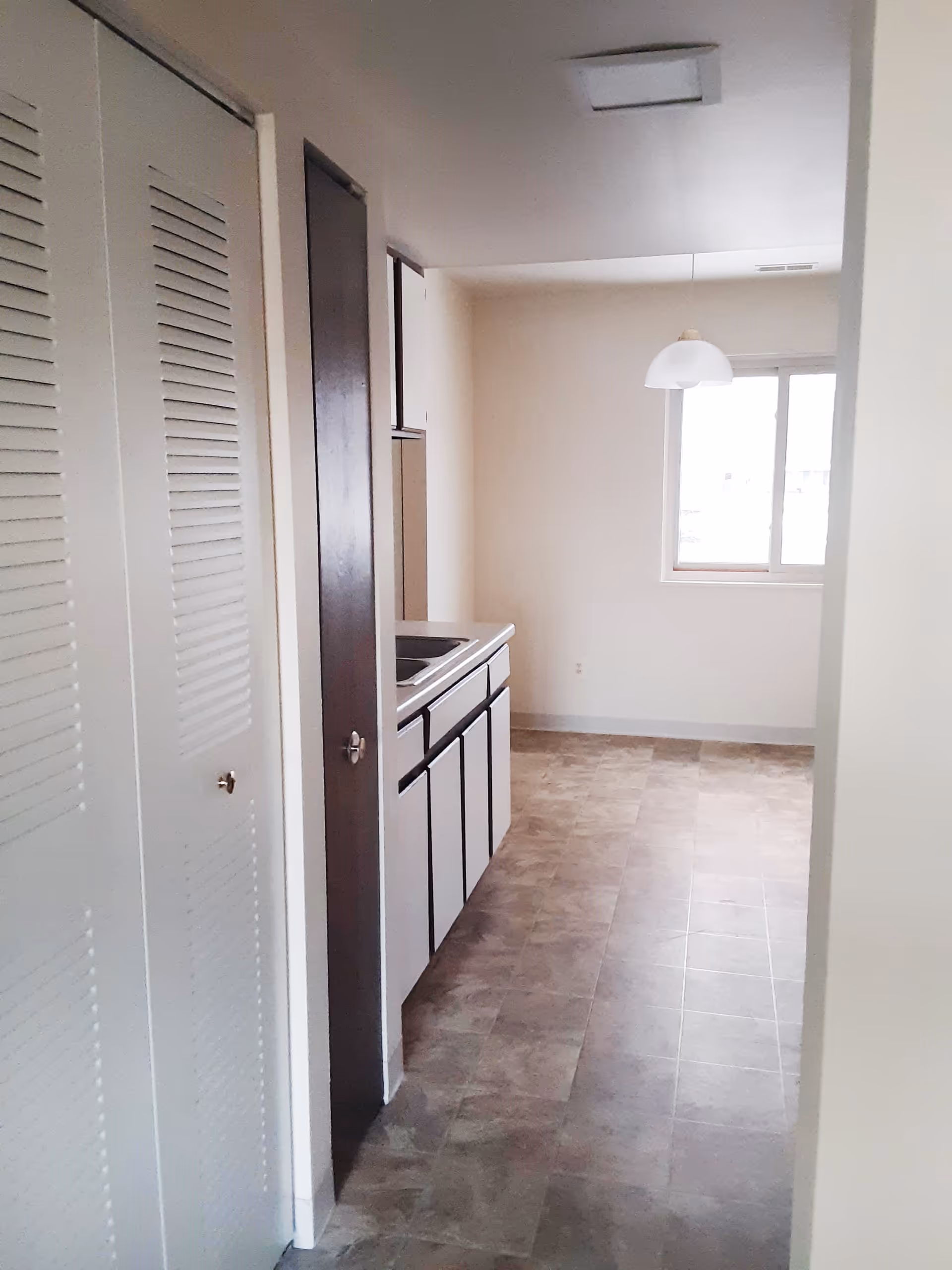 Interior view of a small kitchen area with a sink and cabinets on the left side, a window at the far end, and a hanging light fixture above an empty floor space. The walls are light-colored and the floor has a tiled pattern.