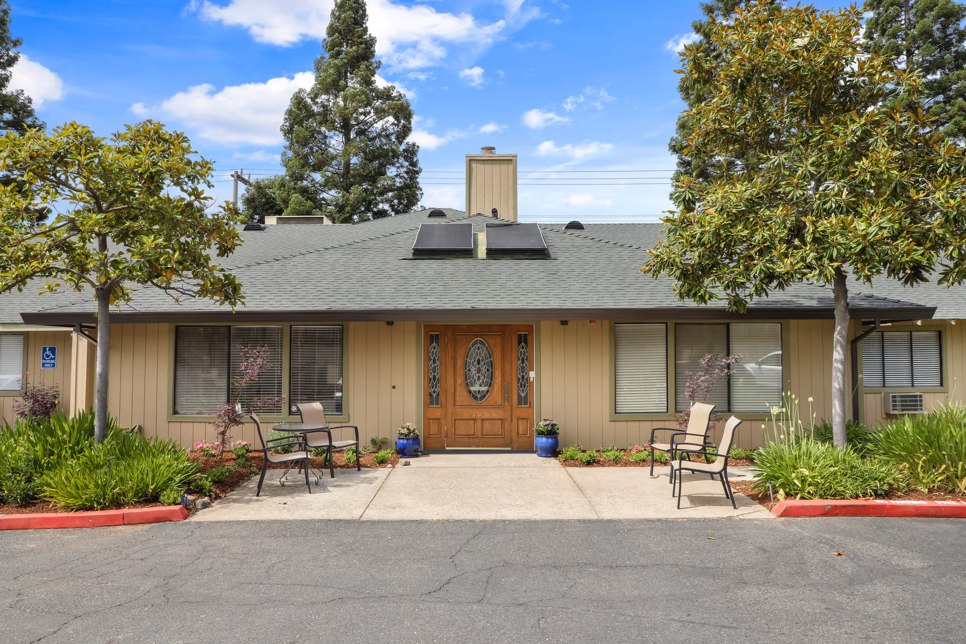 Front exterior view of Almond Grove Assisted Living facility showing a single-story building with a gray shingled roof, wooden double front doors with decorative glass panels, two large windows on either side, and a small seating area with chairs and tables. The entrance is flanked by green shrubs, small trees, and potted plants under a partly cloudy blue sky.