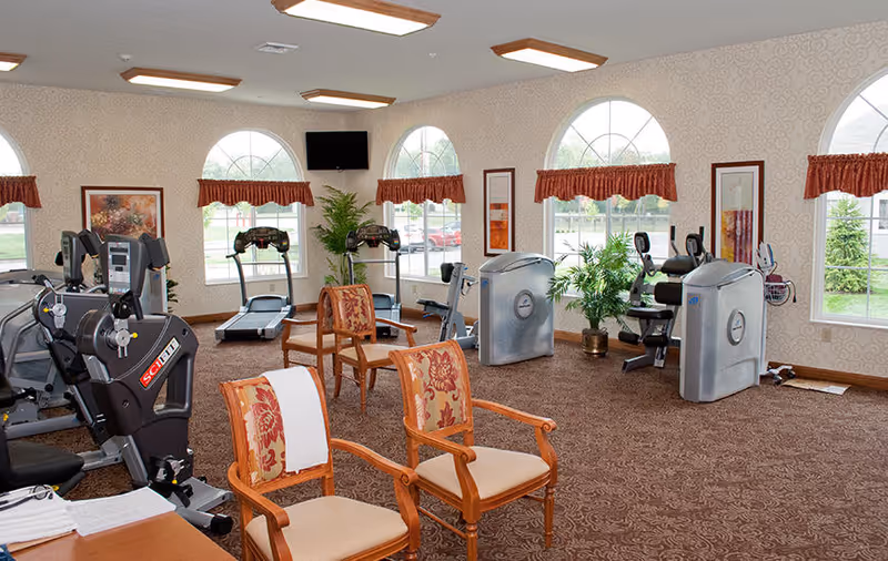 A fitness room with exercise equipment including treadmills, stationary bikes, and weight machines. The room has patterned carpet, several wooden chairs with floral cushions, large arched windows with red valances, framed artwork on the walls, and a wall-mounted TV.