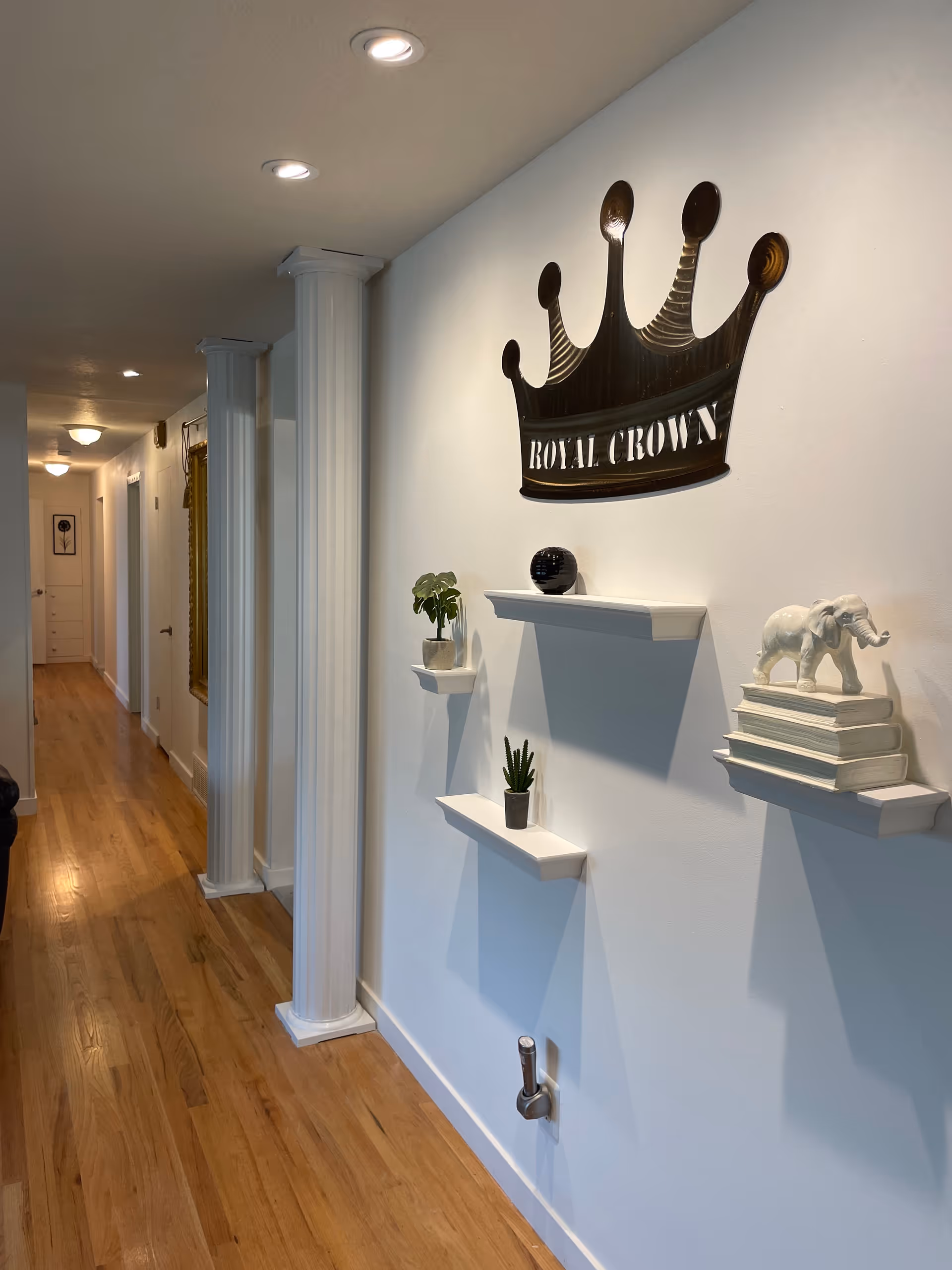 Bright hallway with hardwood floors, decorative white columns and wall shelves beneath a large metal crown sign that reads "Royal Crown".
