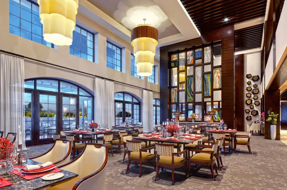 Elegant dining room with multiple tables set with red napkins, plates, glasses, and floral centerpieces. The room features high ceilings with large yellow pendant lights, tall windows with white curtains, and a decorative wall with colorful abstract panels. There are cushioned chairs around the tables and an outdoor patio visible through glass doors.