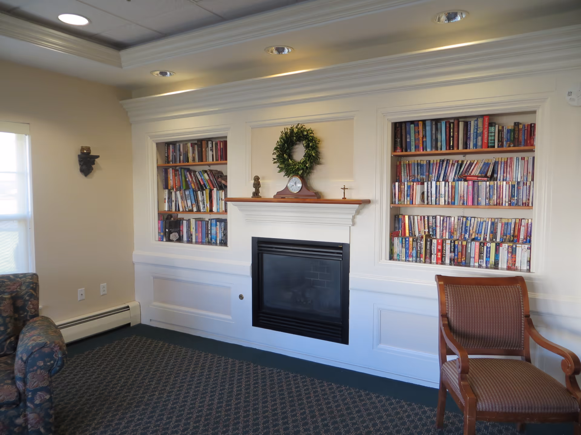 A cozy room with a fireplace centered between two built-in bookshelves filled with books. Above the fireplace mantel is a small clock and a green wreath. To the left is a floral patterned armchair and to the right is a wooden chair with a cushioned seat. The room has beige walls, a patterned carpet, and recessed ceiling lights.