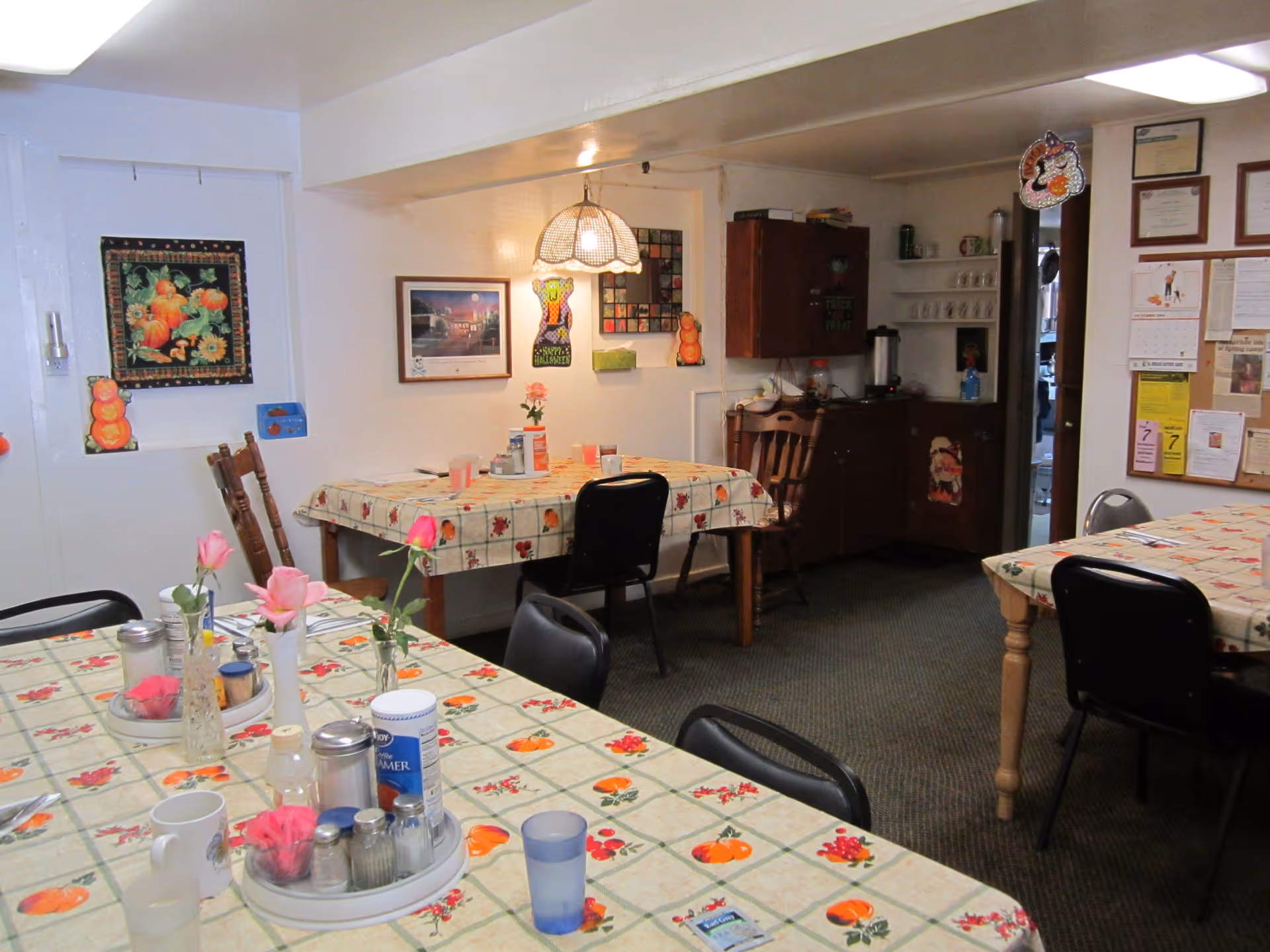 Interior view of a dining room with tables covered in tablecloths featuring a pumpkin and berry pattern. The tables have various items including flowers in vases, cups, salt and pepper shakers, and creamers. The room has chairs around the tables, wall decorations including framed pictures and seasonal decorations, and a bulletin board with papers pinned on it. There is a cabinet and shelves with cups in the background.