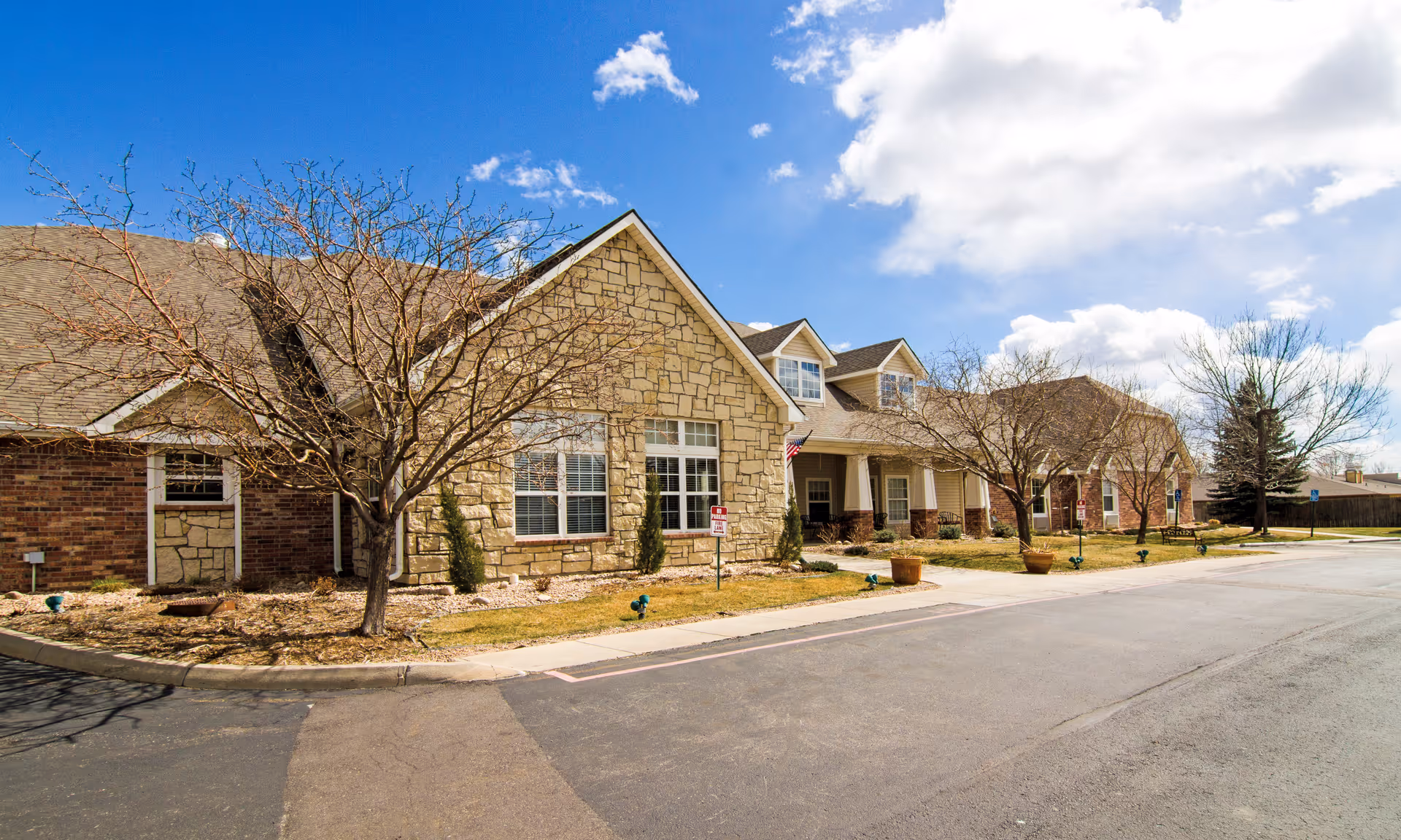Front exterior of a single-story stone-and-brick senior living building with a driveway, leafless trees, and a partly cloudy sky.