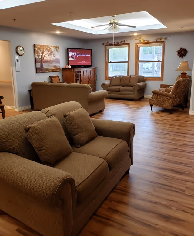 A cozy living room in a senior living facility with multiple cushioned sofas and armchairs arranged around a wooden cabinet holding a flat-screen TV. The room has wooden flooring, two windows with wooden frames, a ceiling fan with lights, a wall clock, a painting of a tree, and a table lamp on a side table.