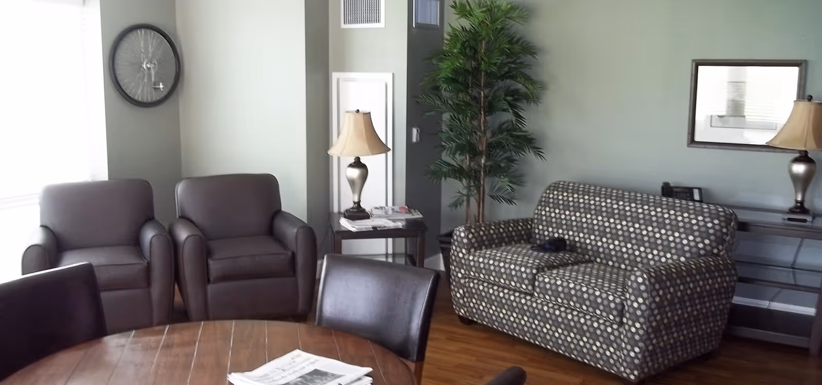 A cozy living room area with a patterned loveseat, two dark armchairs, a round wooden table with newspapers on it, two table lamps on side tables, a tall green plant, a wall mirror, and a bicycle wheel mounted on the wall near a window with blinds.