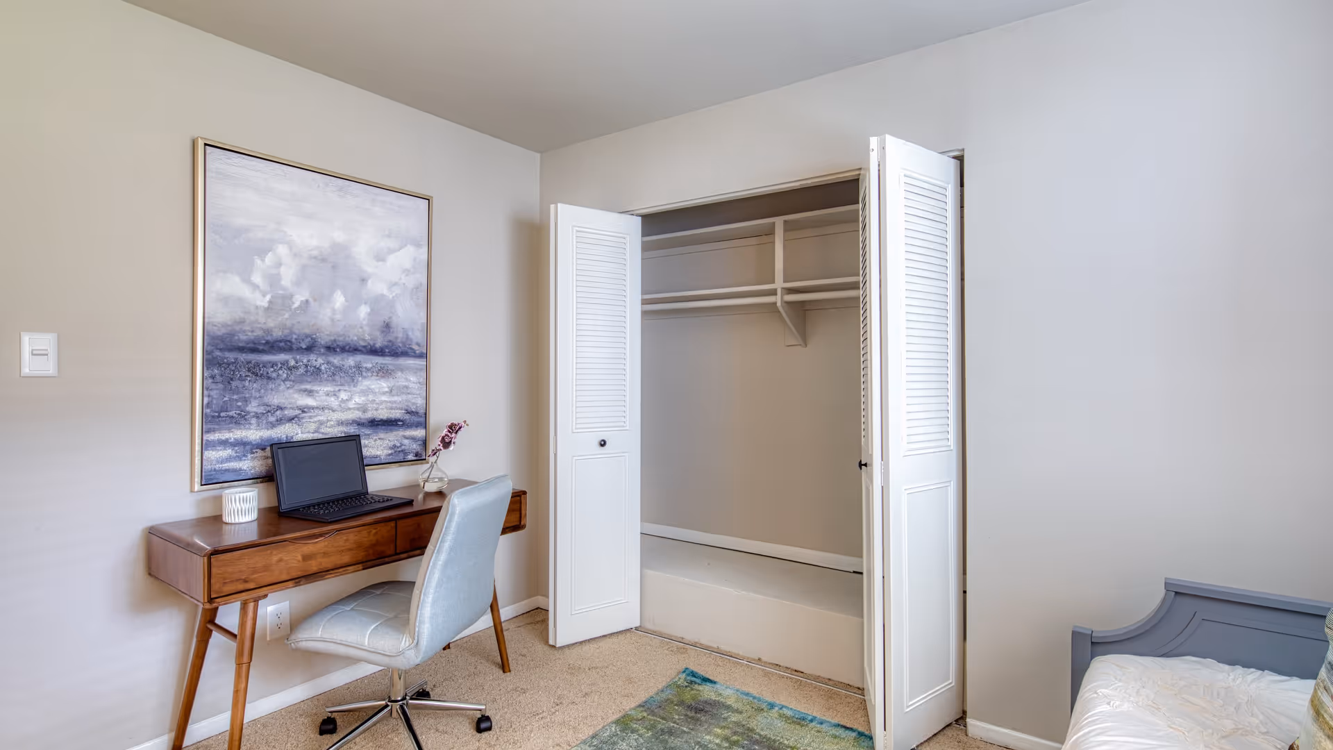 A small bedroom corner featuring a wooden desk with a laptop, a light blue cushioned office chair, a large framed painting of a cloudy sky above water on the wall, and an open closet with white bi-fold doors. Part of a bed with a white comforter and a blue-gray headboard is visible on the right side.