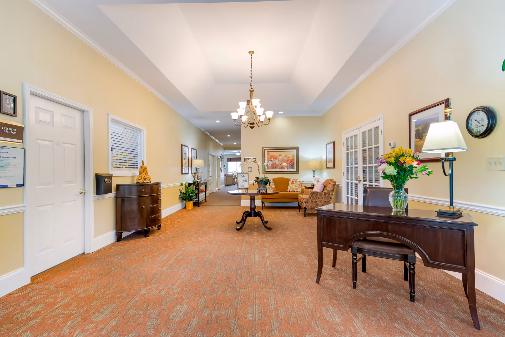 A well-lit hallway in a senior living facility with beige walls and patterned carpet. On the right side, there is a dark wooden desk with a vase of colorful flowers and a lamp. Further back, there is a seating area with a mustard yellow sofa and a patterned armchair, accompanied by framed artwork and lamps. On the left side, there is a door labeled 'Executive Director' and a small dark wooden cabinet with decorative items on top. A chandelier hangs from the ceiling, illuminating the space.