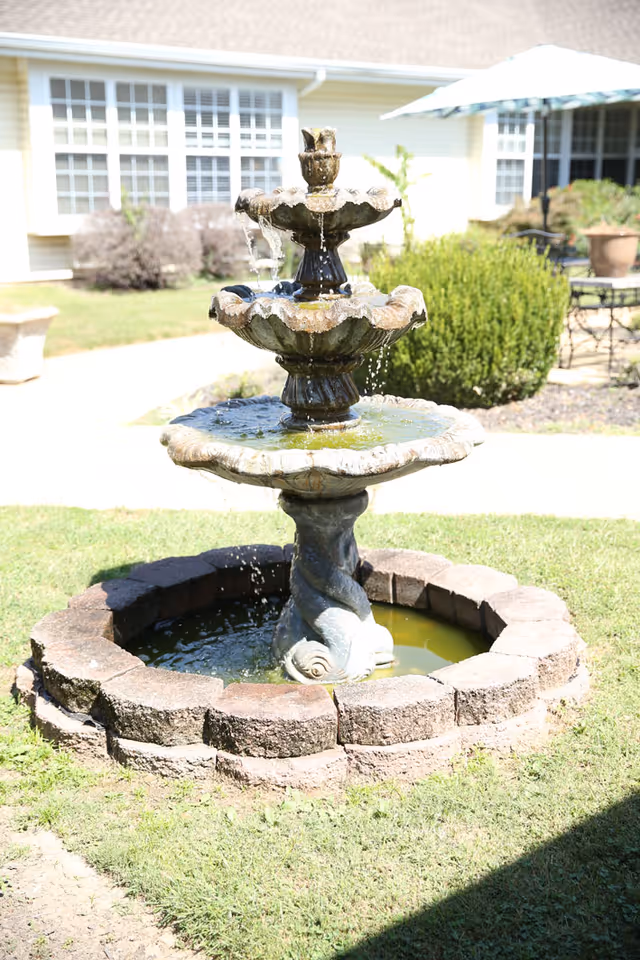 A three-tiered stone water fountain with water flowing from the top tier to the bottom, surrounded by a circular brick border. The fountain is situated on a grassy area with a building, bushes, and outdoor furniture visible in the background.