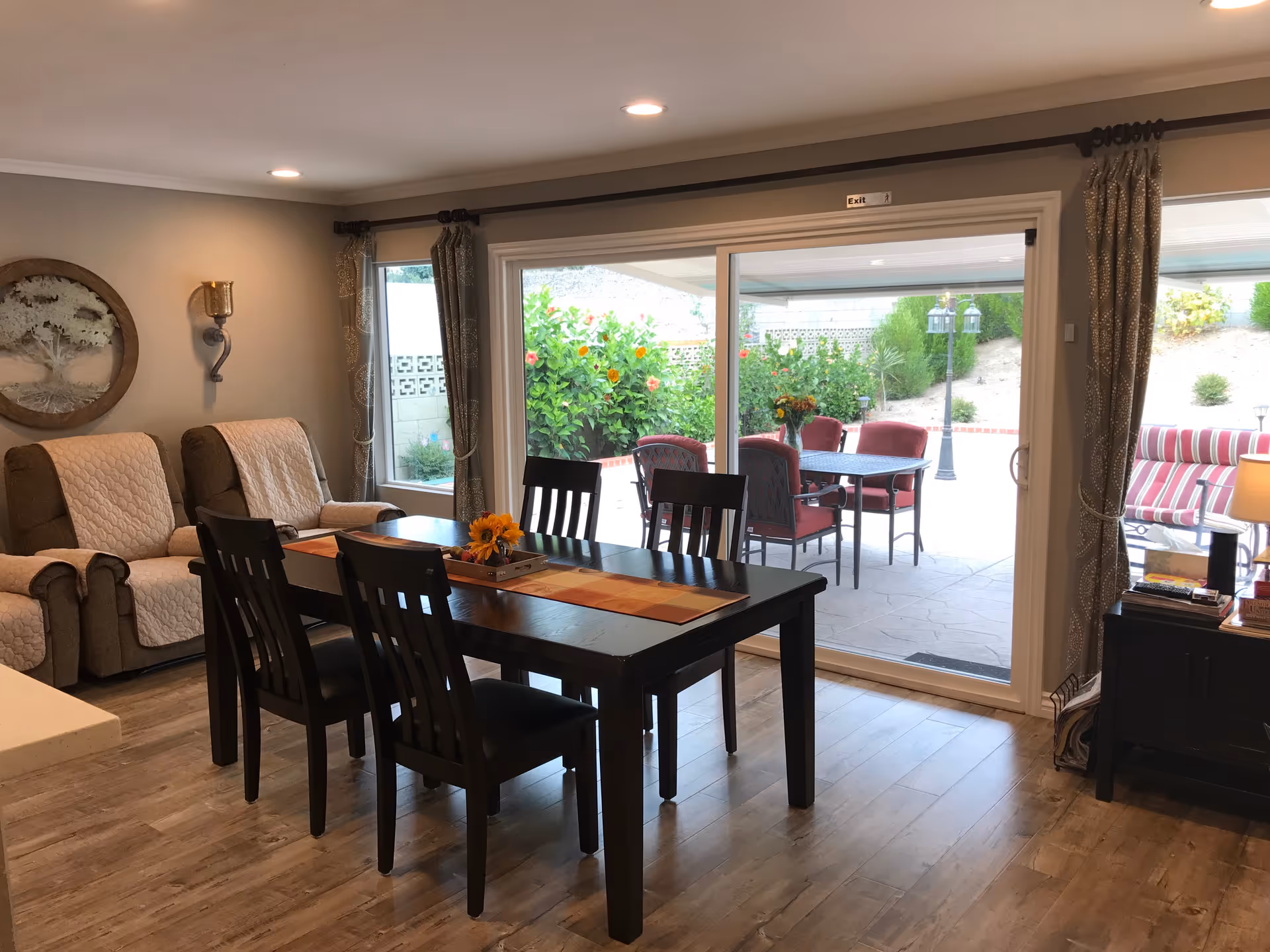 Interior view of a senior living facility dining area with a dark wooden table and four chairs. Behind the table, there are two beige recliners with quilted covers. Large sliding glass doors open to an outdoor patio with red cushioned chairs and a table, surrounded by greenery and flowers. The room has wood flooring, beige walls, and decorative curtains on the windows.