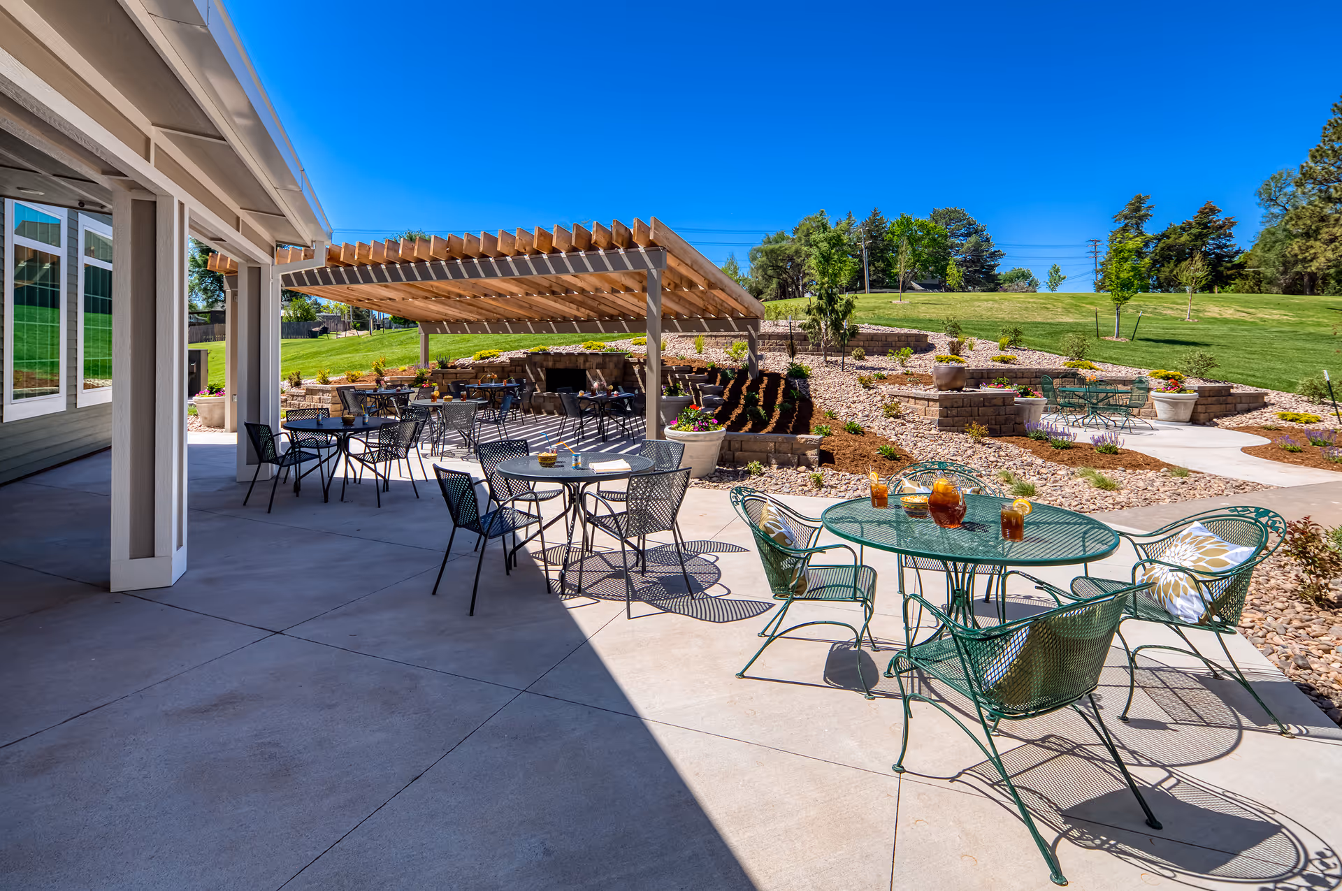 Outdoor patio area with multiple metal tables and chairs, some with drinks on them, under a wooden pergola and open sky. The patio is adjacent to a building with large windows, and there is a landscaped garden with grass, plants, and stone pathways in the background.