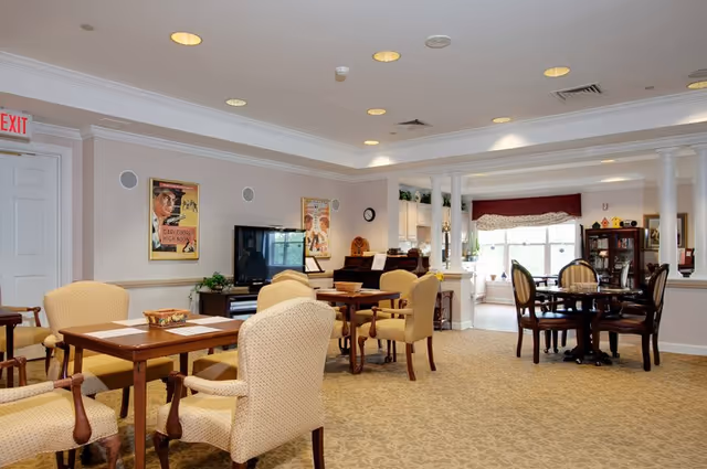 A well-lit common area in a senior living facility featuring multiple tables with upholstered chairs arranged for socializing or activities. The room has beige carpeting, light-colored walls with framed artwork, a television on a stand, and a piano. Large windows in the background allow natural light to enter, and decorative columns partially separate the space.