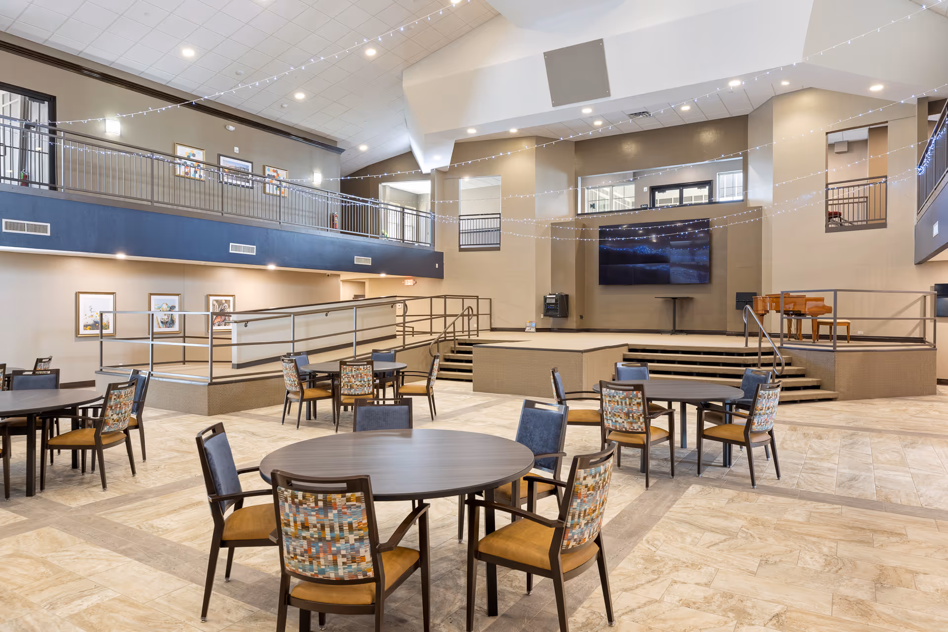 Spacious common area with round tables and chairs arranged on a tiled floor, a raised stage with a piano and large TV screen mounted on the wall, string lights hanging from the ceiling, and a second-floor balcony overlooking the space.