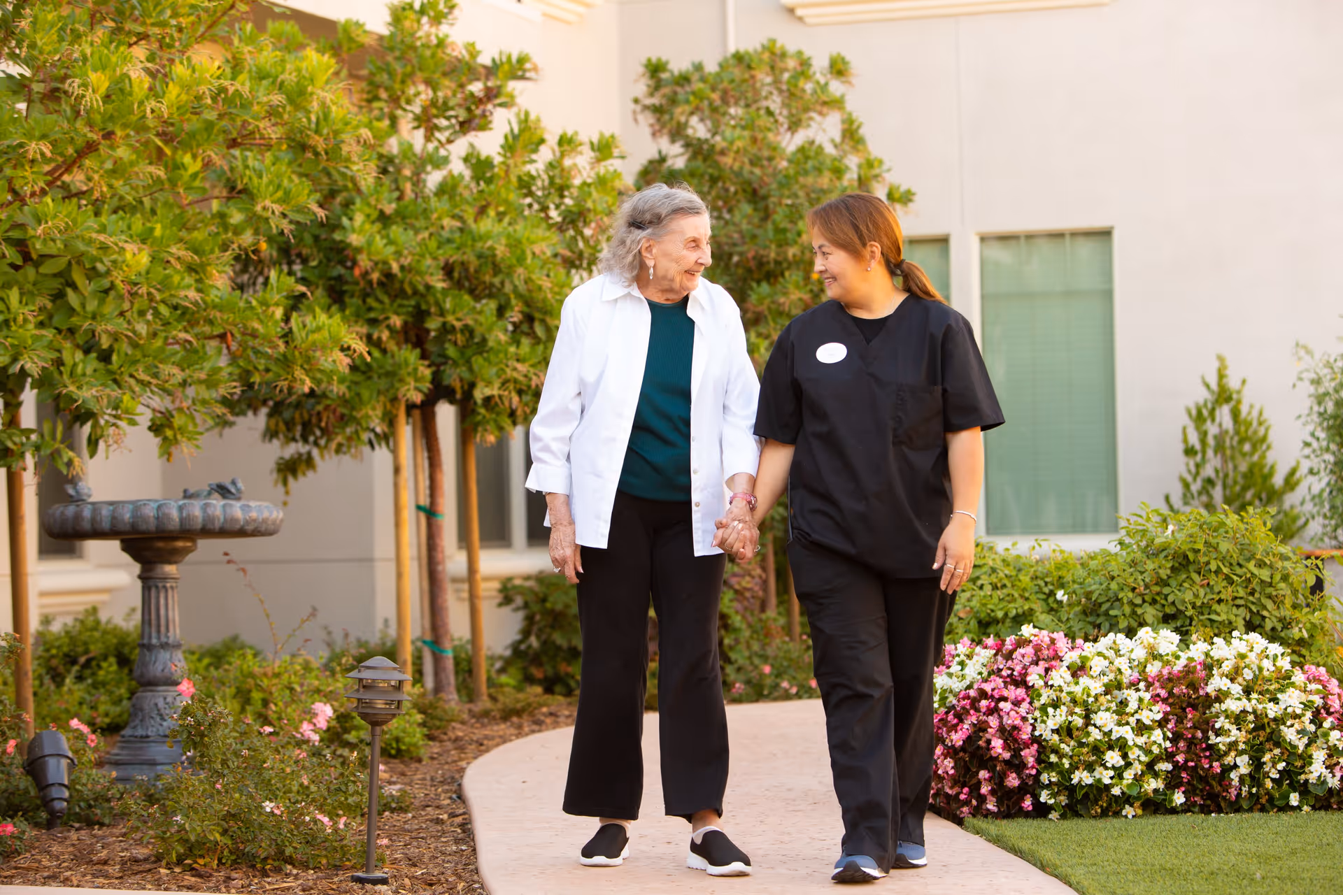 An elderly woman and a caregiver holding hands and walking along a garden path outside a building. The garden is well-maintained with green bushes, flowering plants, and small trees. The caregiver is wearing black scrubs and the elderly woman is dressed in a white jacket and black pants. Both are smiling and looking at each other.