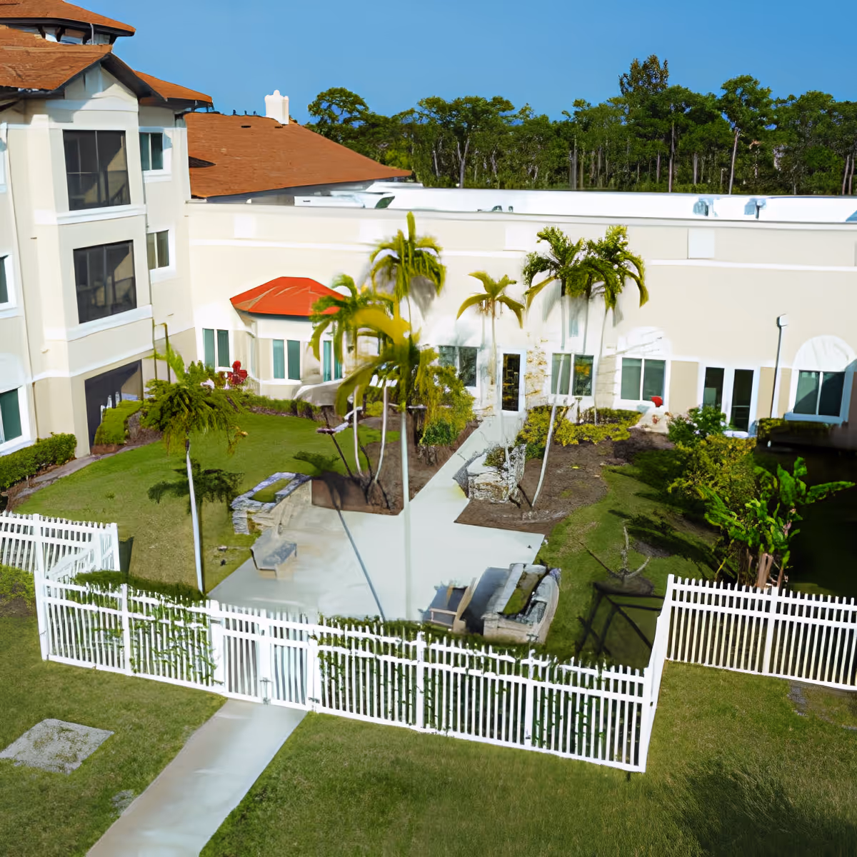 A landscaped fenced courtyard with palm trees, walkways, and seating beside a multi-story senior living building.