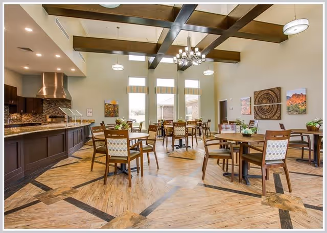 A spacious dining room with multiple round wooden tables and chairs arranged neatly. The room features large windows allowing natural light to fill the space, wooden ceiling beams, modern light fixtures, and a kitchen area with a stainless steel range hood and dark cabinetry.