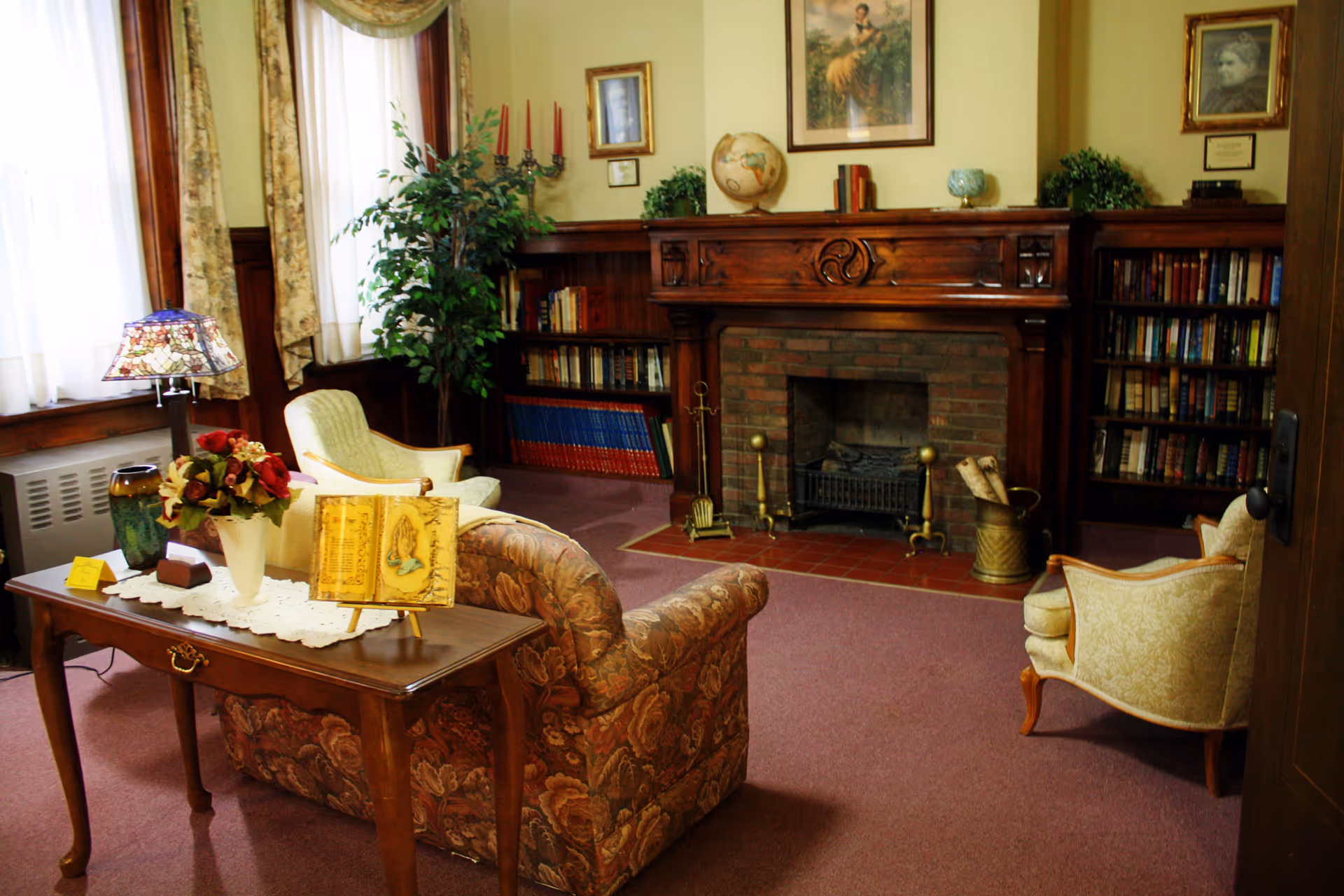 A cozy living room with vintage-style furniture including a floral-patterned sofa, two armchairs, and a wooden table with a lace runner, flowers, and a decorative book. The room features a wooden fireplace with a brick interior, flanked by bookshelves filled with books. There are framed pictures and a globe on the mantel, a potted plant near the window, and curtains allowing natural light to enter.