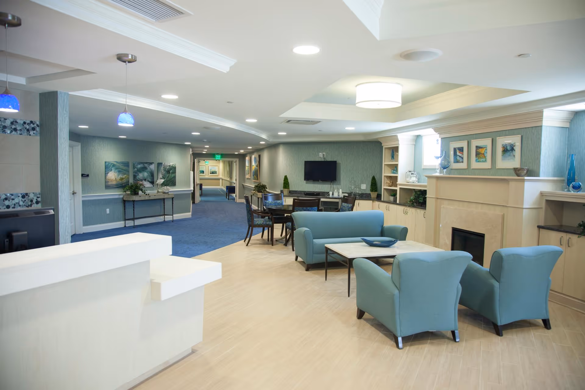 A spacious and well-lit common area in a senior living facility featuring light blue armchairs and a sofa arranged around a coffee table in front of a fireplace. The room has light wood flooring, built-in shelves with decorative items, framed artwork above the fireplace, and a flat-screen TV mounted on the wall. A hallway with blue carpeting and additional seating is visible in the background.