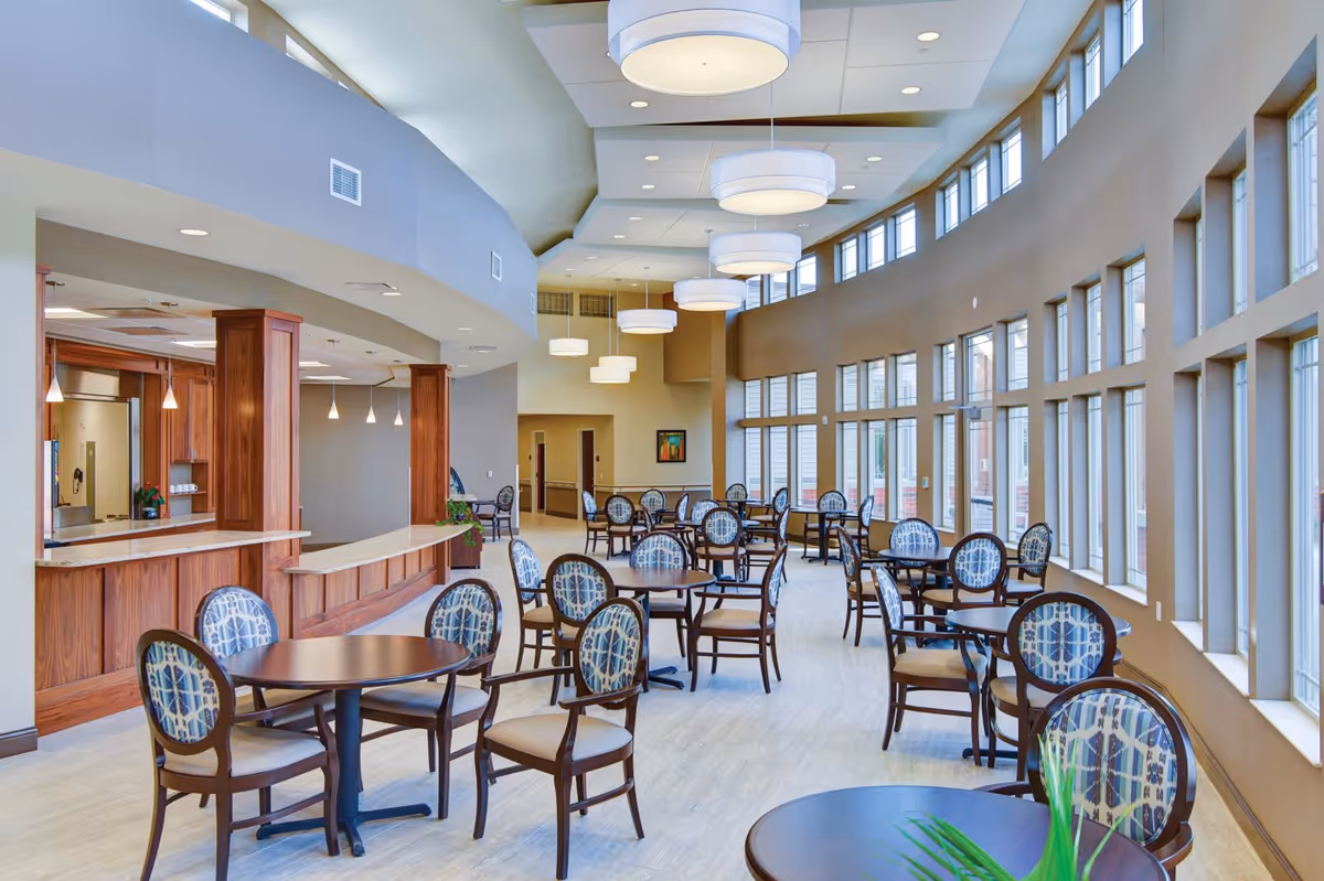 A spacious dining room with multiple round tables and chairs featuring patterned upholstery. The room has large windows along one wall allowing natural light to fill the space. There are modern hanging light fixtures from the ceiling and a serving counter with wooden cabinetry on the left side.