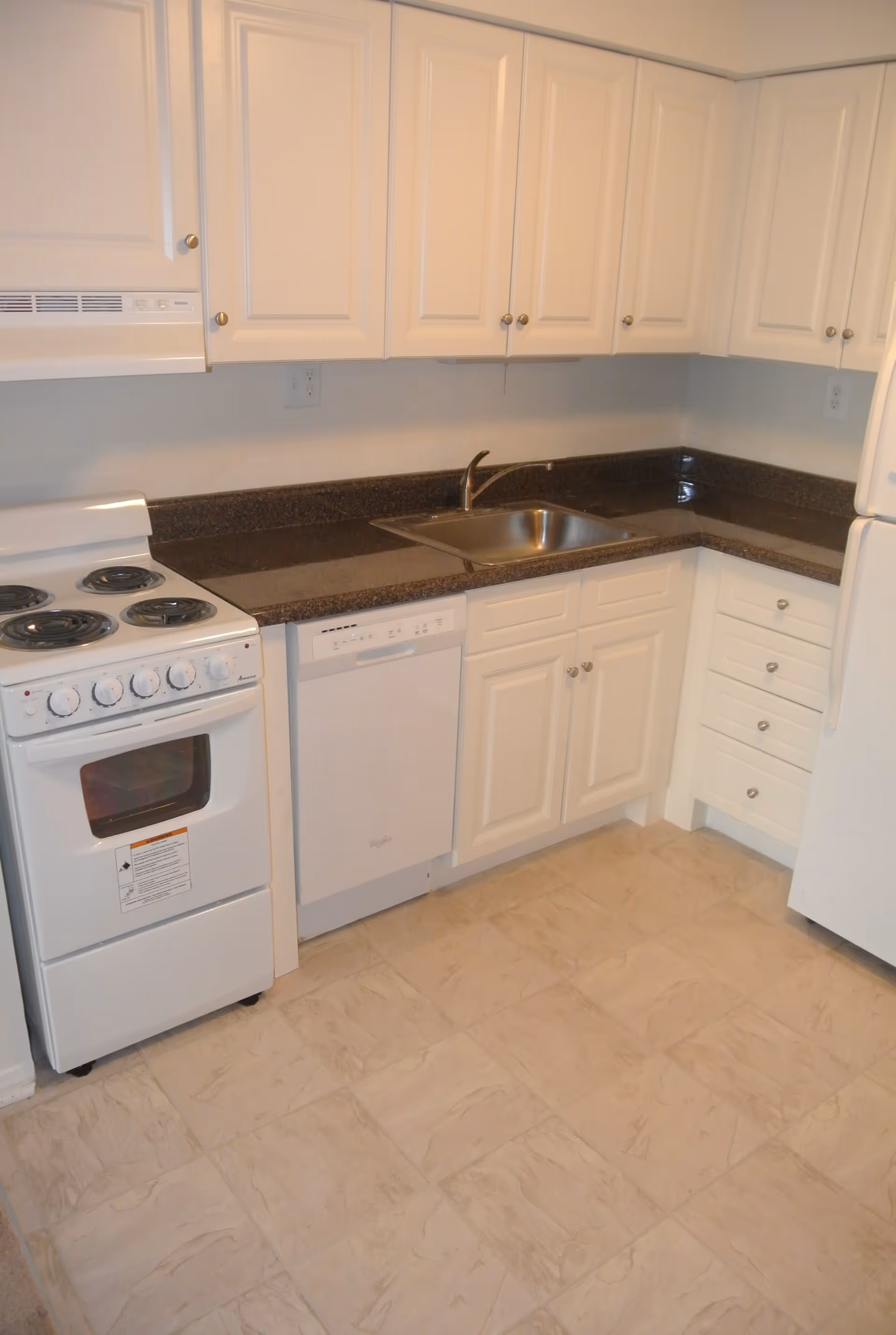 A clean kitchen with white cabinets, a white electric stove with four burners, a white dishwasher, a stainless steel sink with a faucet, and a white refrigerator. The countertops are dark brown, and the floor has light-colored tiles.