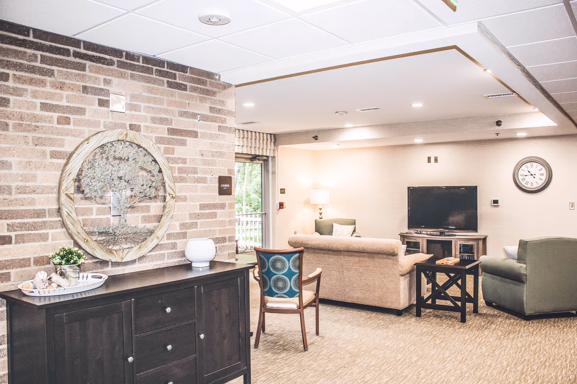 A cozy lounge area in a senior living facility with a beige couch, green armchair, wooden side table, and a flat-screen TV mounted on a wooden stand. The room has a brick accent wall with a decorative tree artwork and a dark wooden cabinet with decorative items on top. A clock is mounted on the wall above the TV, and a lamp provides warm lighting in the corner.