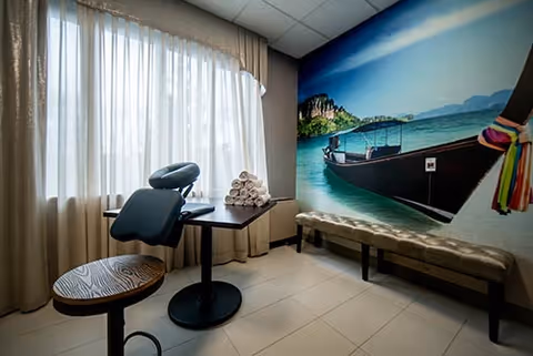 A small room with a massage chair, a round wooden stool, a table with a stack of rolled white towels, and a bench along the wall. One wall features a large mural of a boat on turquoise water with a scenic background of cliffs and sky. Light filters in through sheer curtains covering a window.