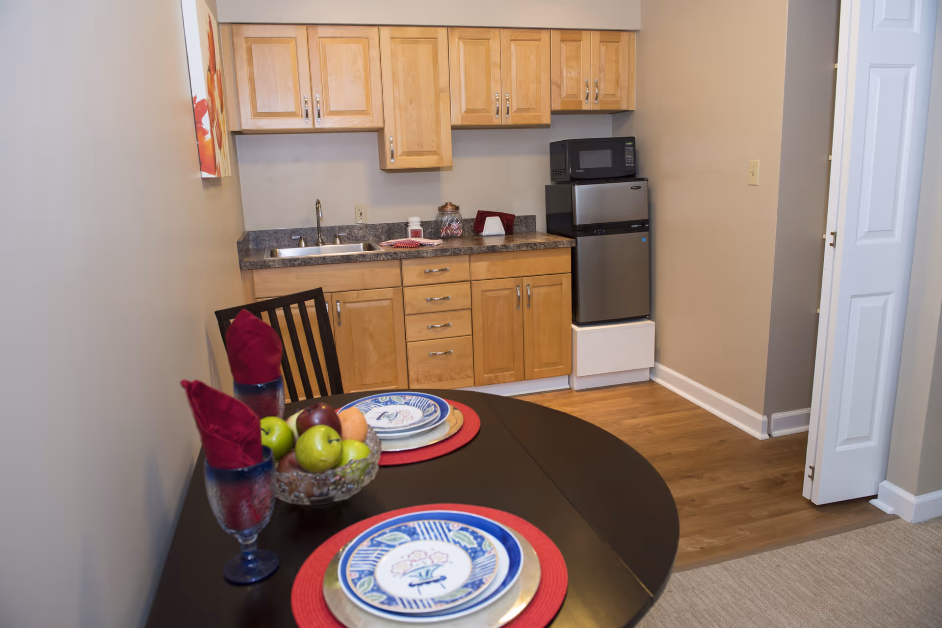 A small kitchen area with wooden cabinets, a countertop with a sink, a microwave on top of a mini refrigerator, and a dining table set for two with plates, glasses, and a bowl of fruit.