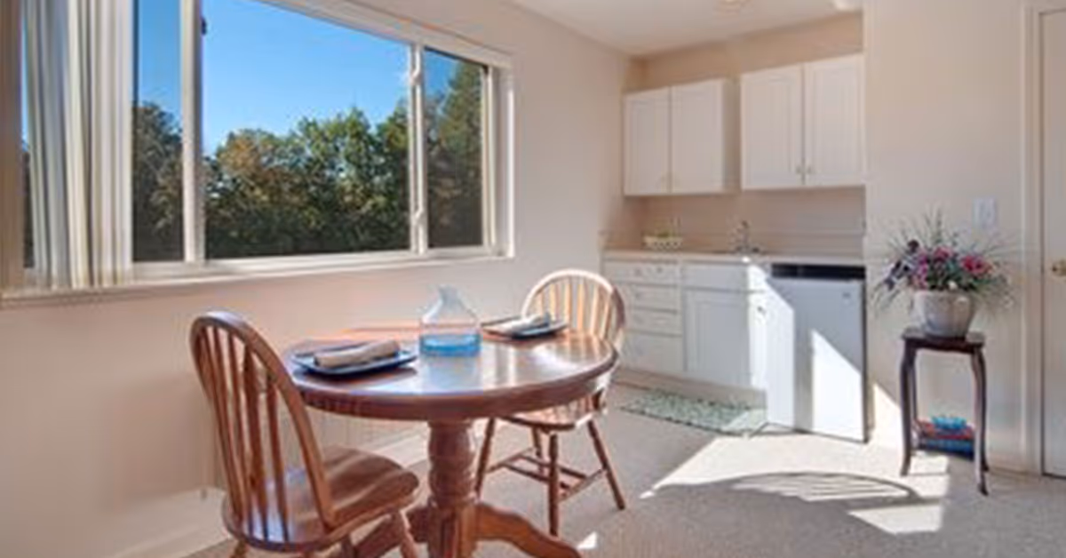 A bright kitchen area with white cabinets, a small refrigerator, and a round wooden table set with two wooden chairs. A large window shows green trees outside, and a small table with a flower arrangement is placed near the door.