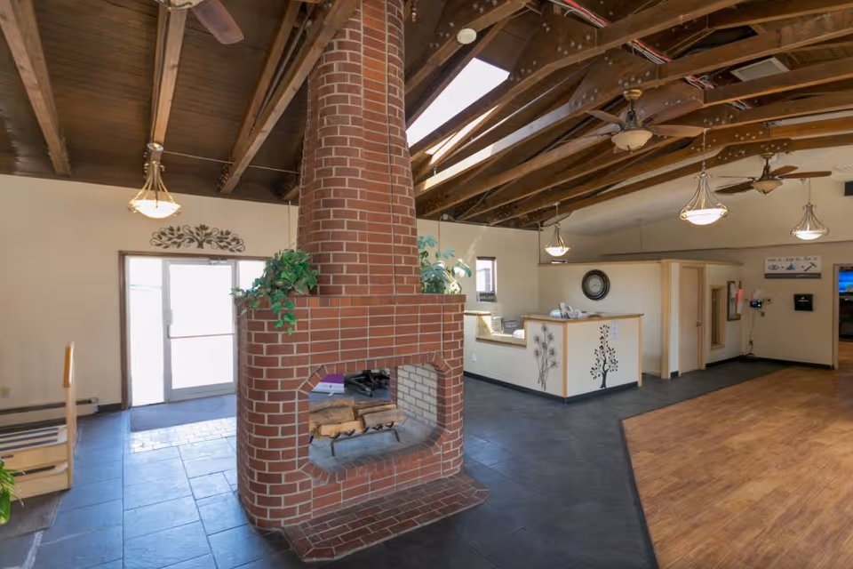 Interior view of a senior living facility lobby with a central brick fireplace, wooden ceiling beams with ceiling fans and hanging lights, a reception desk decorated with tree designs, and a glass door entrance letting in natural light.