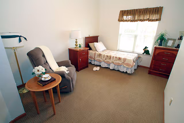 A simple bedroom with a single bed covered with a quilt, a wooden nightstand with a lamp, a wooden dresser, a window with a brown valance, a gray recliner chair with a white throw blanket, and a small round wooden side table with a flower vase and a teacup set.