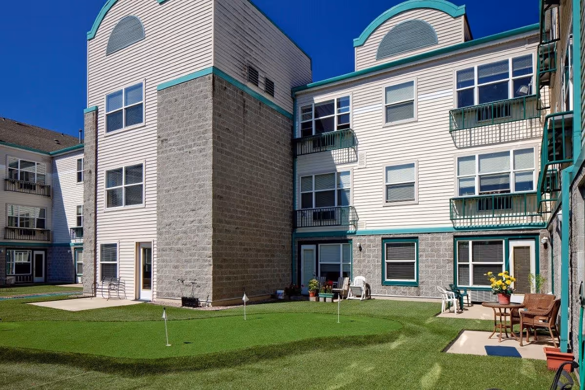 Exterior view of Hazelwood Community Apartments showing a courtyard with a small putting green, surrounded by a three-story building with multiple windows and balconies. There are chairs, tables, and potted plants arranged on the concrete patios around the green. The sky is clear and blue.