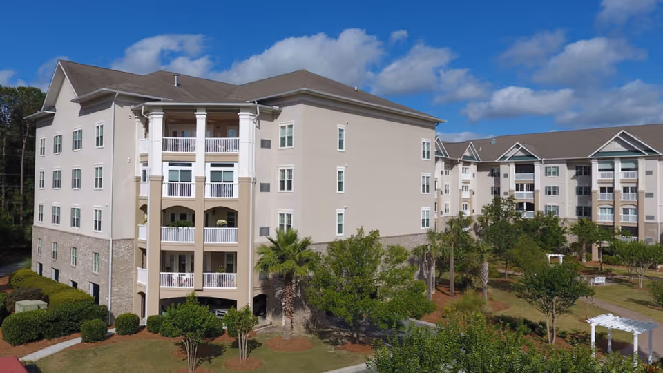 Front exterior of a multi-story senior living building with balconies, landscaped grounds and palm trees under a blue sky.