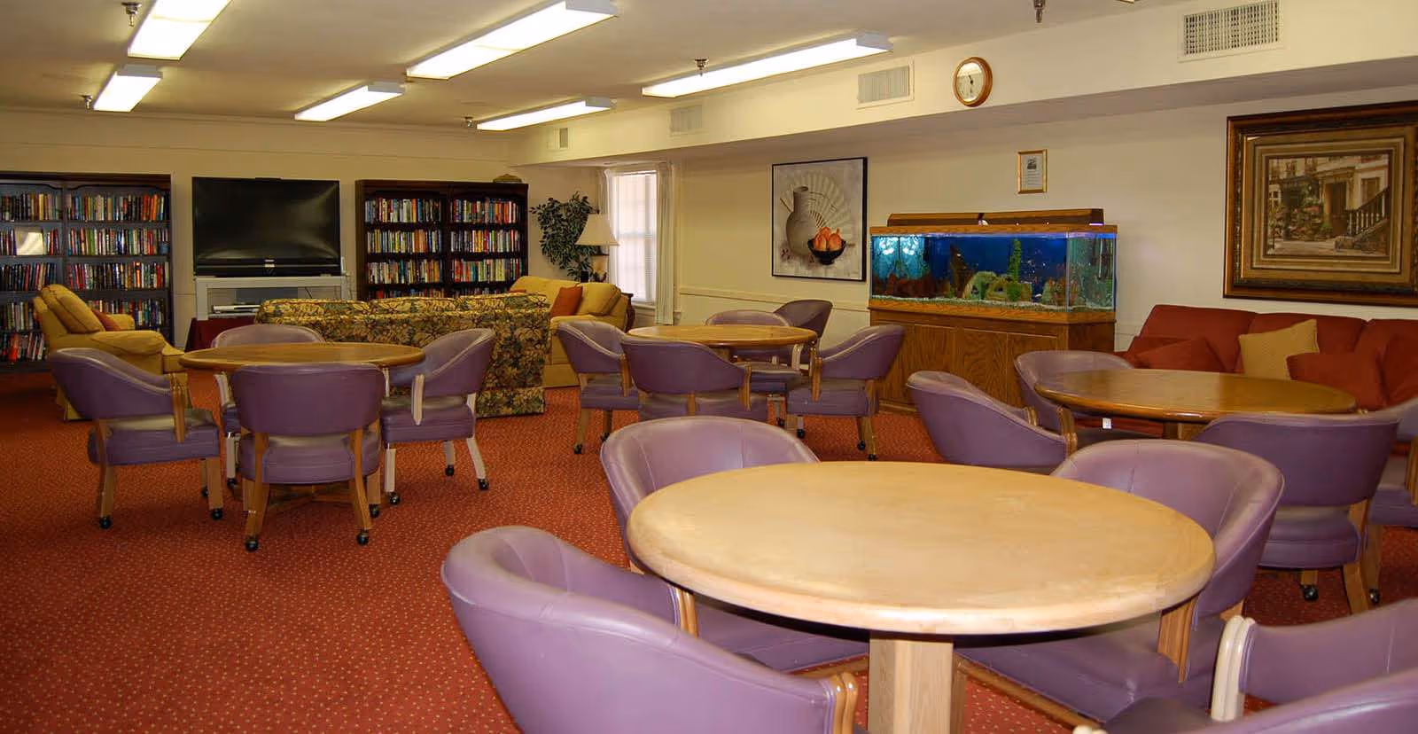 A senior living facility common area with multiple round tables surrounded by purple cushioned chairs. In the background, there are bookshelves filled with books, a large flat-screen TV, a floral patterned sofa, a fish tank on a wooden stand, and framed artwork on the walls. The room has a red carpet and fluorescent ceiling lights.