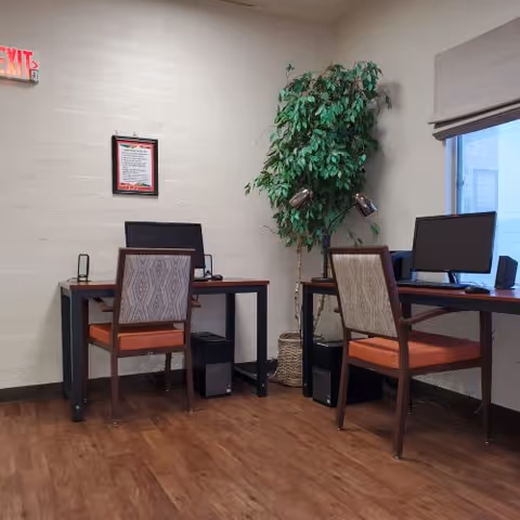 A small office area with two wooden desks, each with a computer monitor and chair with orange cushions. There is a tall green potted plant between the desks, a framed document on the wall, and a window with a beige blind. The floor is wooden, and an exit sign is visible above a door on the left.