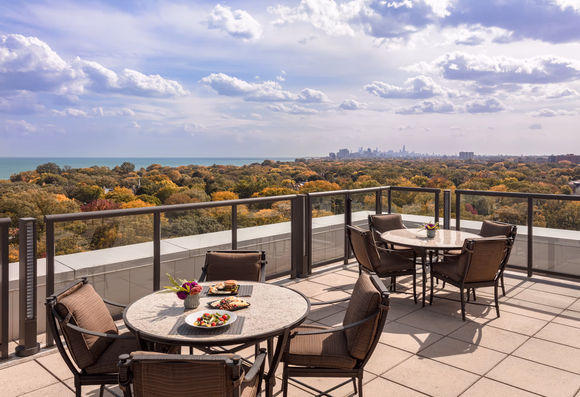 Outdoor terrace with two round tables and several cushioned chairs, overlooking a scenic view of a forested area with autumn foliage and a distant city skyline under a partly cloudy sky.