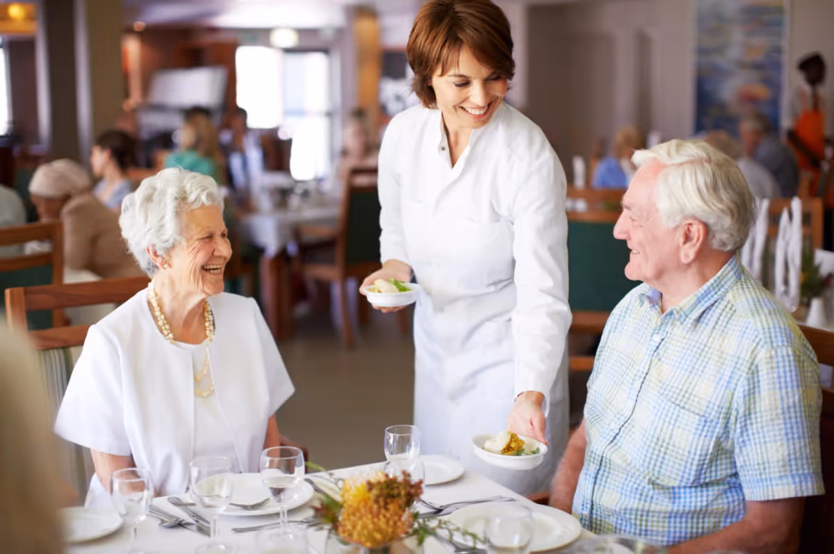 A smiling female server in a white uniform serves food to an elderly man and woman seated at a dining table in a senior living facility dining room. The elderly woman is wearing a white blouse and pearl necklace, and the elderly man is wearing a light blue plaid shirt. The table is set with plates, glasses, and silverware, and other people are visible in the background.