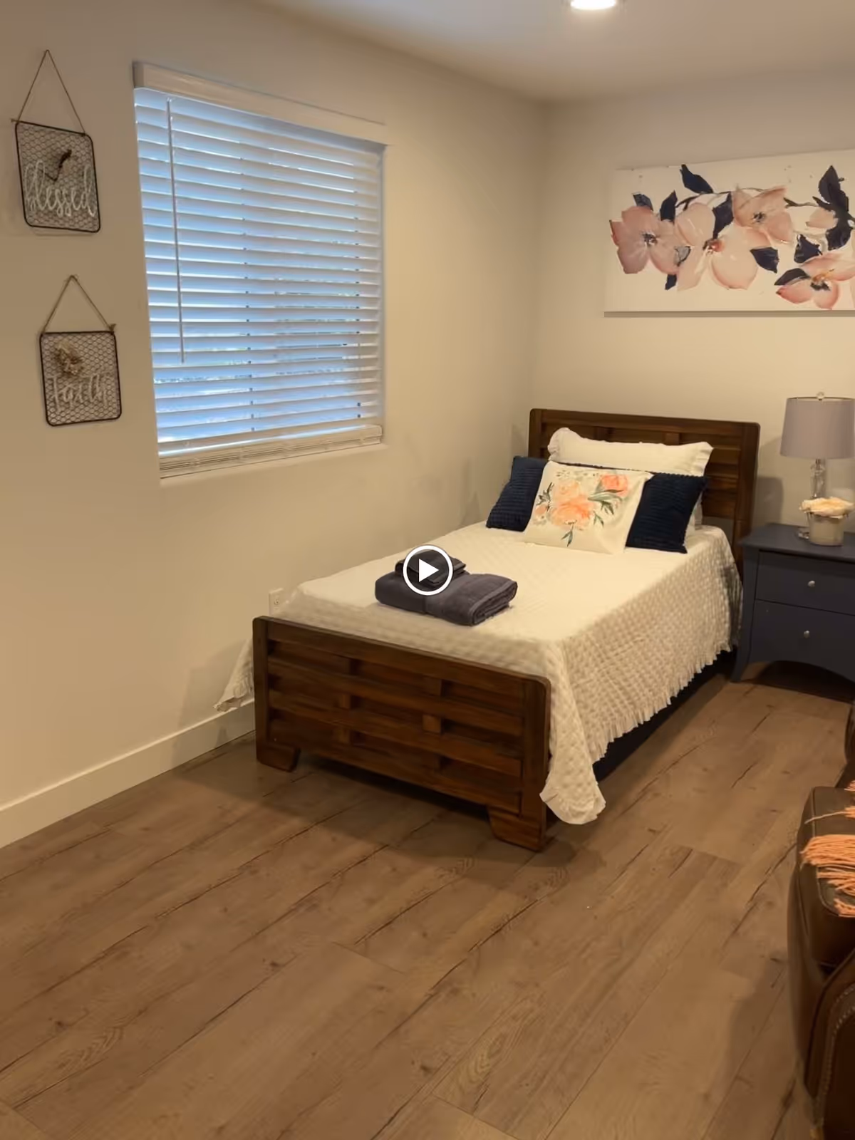 A cozy bedroom with a single wooden bed covered with a white quilt and decorated with floral and dark blue pillows. A folded dark towel is placed on the bed. There is a window with white blinds on the left wall, two decorative wall hangings with the words 'blessed' and 'grateful' beside it. On the right side of the bed, there is a dark blue nightstand with a lamp and a small decorative item. Above the bed, a floral painting with pink flowers is hung on the wall. The floor is wooden with a light brown finish.