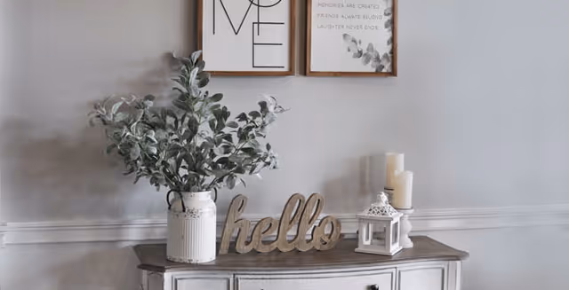 A decorative console table against a light gray wall with two framed prints above it. On the table, there is a white vase with green leafy branches, a wooden decorative sign that says 'hello', a white lantern, and three pillar candles.