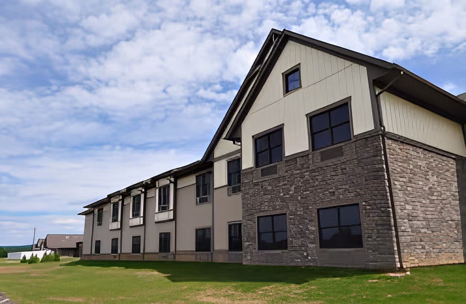 Exterior view of a two-story building with a combination of stone and beige siding under a partly cloudy sky, surrounded by a grassy area.